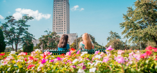Two women sit outside of the Louisiana State Capitol surrounded by plants. 
