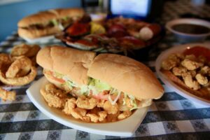A close-up image of a fried shrimp po'boy, with various seafood items on the table behind it.