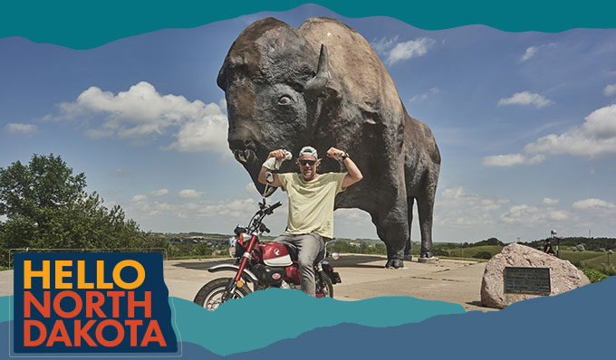 Man sitting on a red motorcycle in front of a large buffalo statue while flexing his arms 