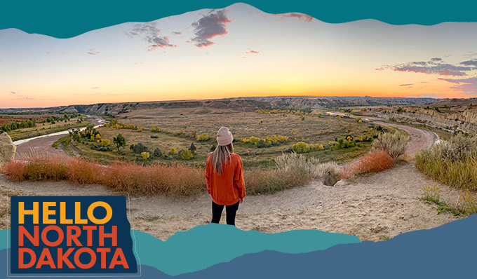 A view of the back of a woman wearing a red sweatshirt looking out to butte landscapes and a sunset