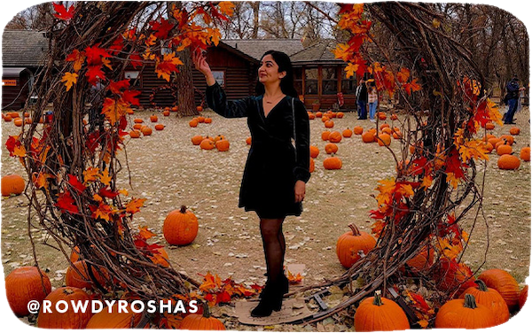 A woman wearing a black dress standing in a large wreath of fall foliage and surrounded by pumpkins, with overlay text that reads @rowdyroshas