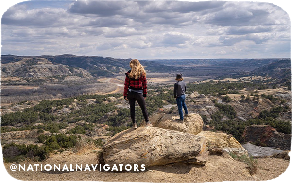 Two people standing on rocks and looking out to hilly terrain with text overlay that reads @NationalNavigators