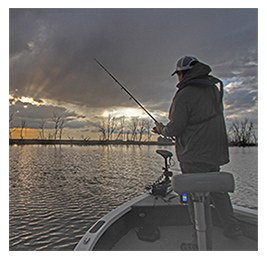 Man standing on a fishing boat casting his fishing rod into water with a sunset behind him