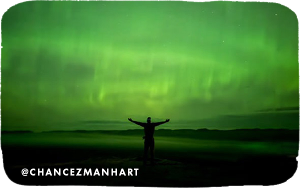 Man standing with his arms outstretched with a green background of the Northern Lights, with overlay text that reads @chancezmanhart