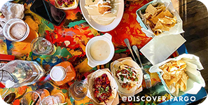 Overhead view of a restaurant table with tacos, chips, cheese dip and drinks on it, with overlay text that reads @discover.fargo