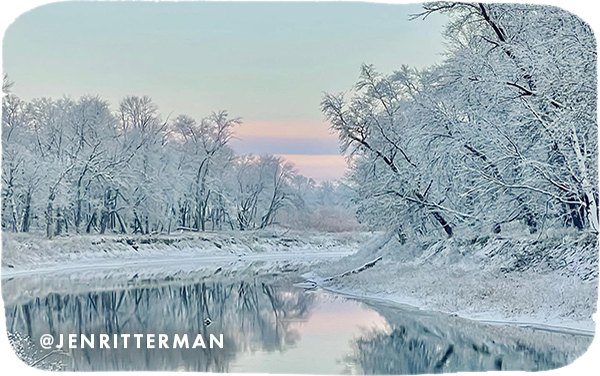 Scene of trees covered in snow reflecting in a glassy surface of water, with overlay text that reads @jenritterman