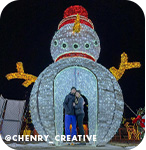 A couple standing in a large snowman holiday light sculpture lit up at night, with overlay text that reads @chenry_creative