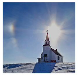 White church building with a red roof sitting on a snowy hill with the sun beaming behind it