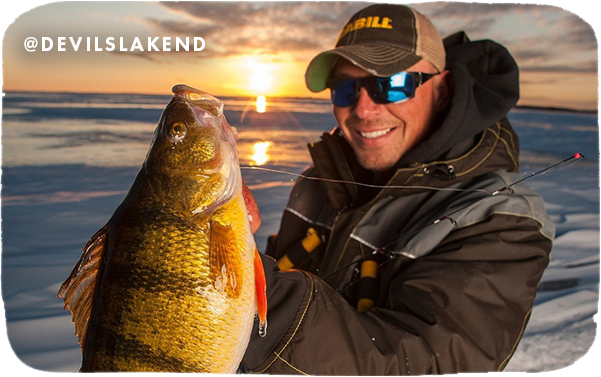 Man wearing a winter jacket and holding a caught fish in front of a frozen lake with a sunset behind him, with text overlay that reads @devilslakend