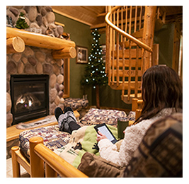 View from behind of a woman reclining in front of a fireplace in a cozy wood cabin