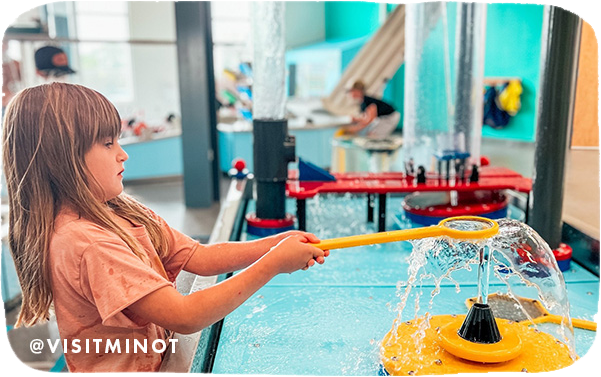 A little girl playing with an interactive water display inside a museum, with overlay text that reads @visitminot