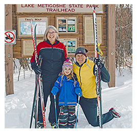 A smiling woman, man and child holding ski equipment and posing in front of a sign that reads: Lake Metigoshe State Park Trailhead