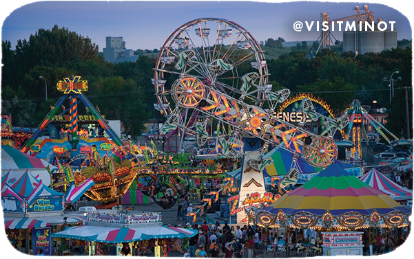 Carnival rides lit up at night, with overlay text that reads @visitminot