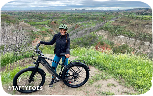 Woman posing with a mountain bike at a vista point overlooking green hills, with overlay text that reads: @tattyfoes