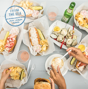 Point of view looking down on a picnic table with lobster rolls, clam rolls, steamed clams, chowder and beer. 