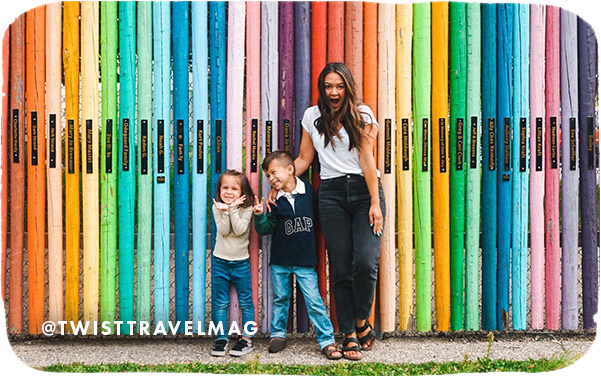 Woman and two children standing in front of colorful wood poles, with overlay text that reads: @twisttravelmag