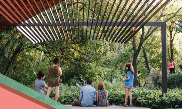 People stand and sit on the ground under a pergola at UMLAUF Sculpture Garden & Museum in Austin, Texas.