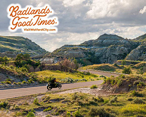 Wide shot of a motorcycle rider on a winding road surrounded by rock formations, with overlay text that reads: Badlands. Good Times. VisitWatfordCity.com
