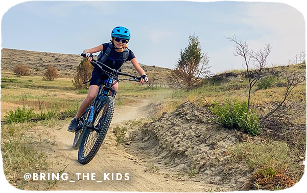 Young child riding a mountain bike on a dirt trail, with overlay text that reads: @bring_the_kids