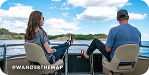 View from behind of a man and a woman sitting on a boat with water and blue sky ahead of them, with overlay text that reads: @wanderthemap
