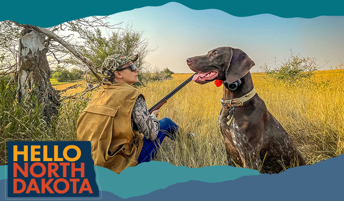A man crouching in tall grass with a dog next to him, with overlay text that reads: Hello North Dakota