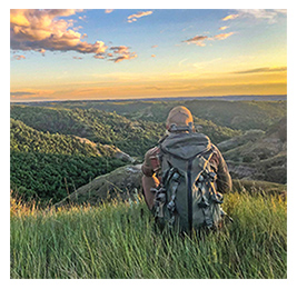 The back of a person sitting in nature with a view of mountains and sunset ahead of them