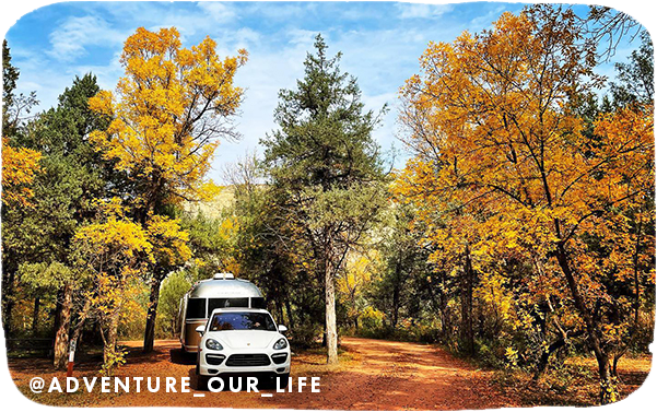 A white car and silver trailer parked and surrounded by trees with gold fall leaves, with overlay text that reads: @adventure_our_life