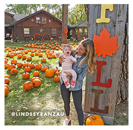 Woman holding a baby at a pumpkin patch with a sign behind her that reads 