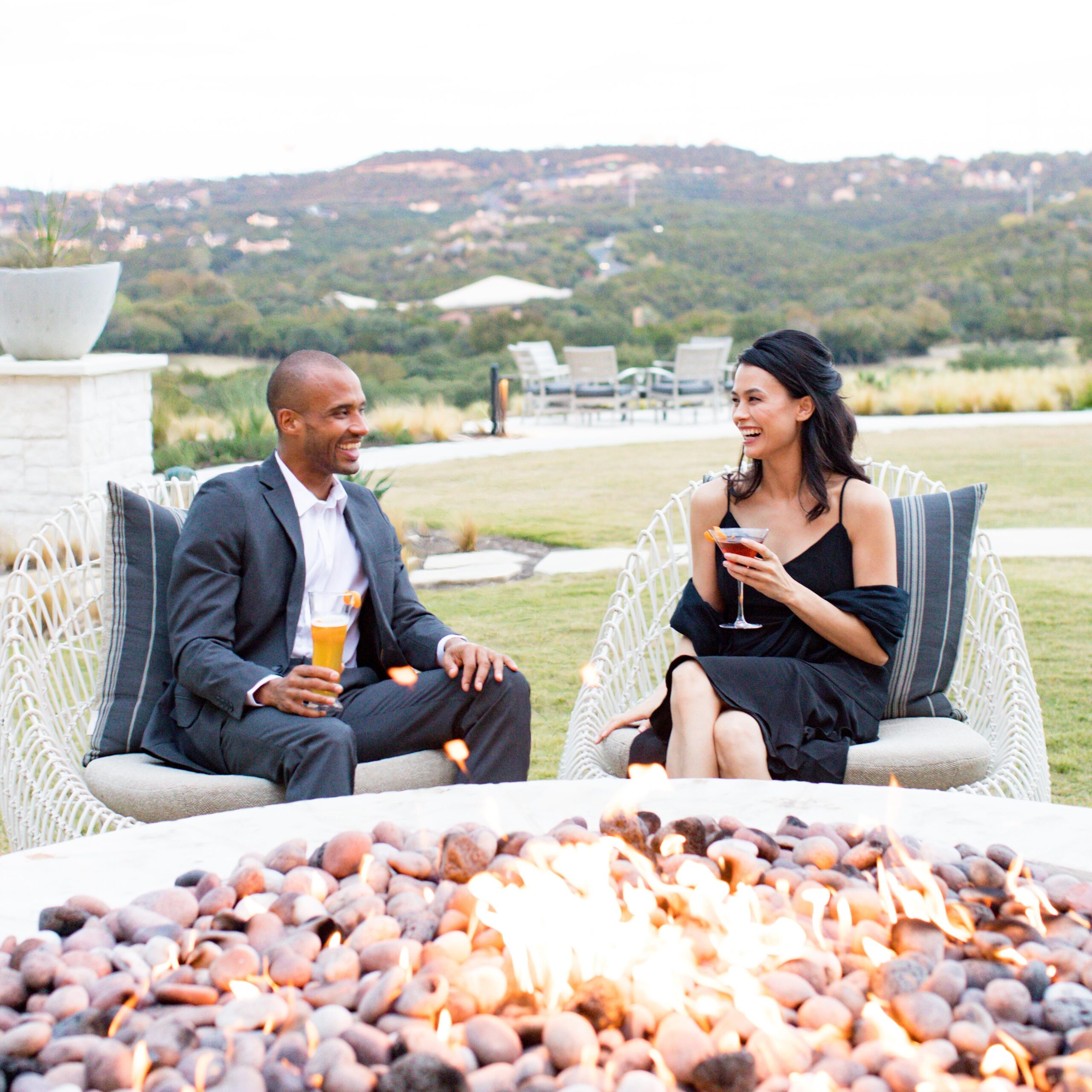 A man and woman enjoys drinks by a fire pit at Austin's Omni Barton Creek Resort & Spa with scenic hills in the background