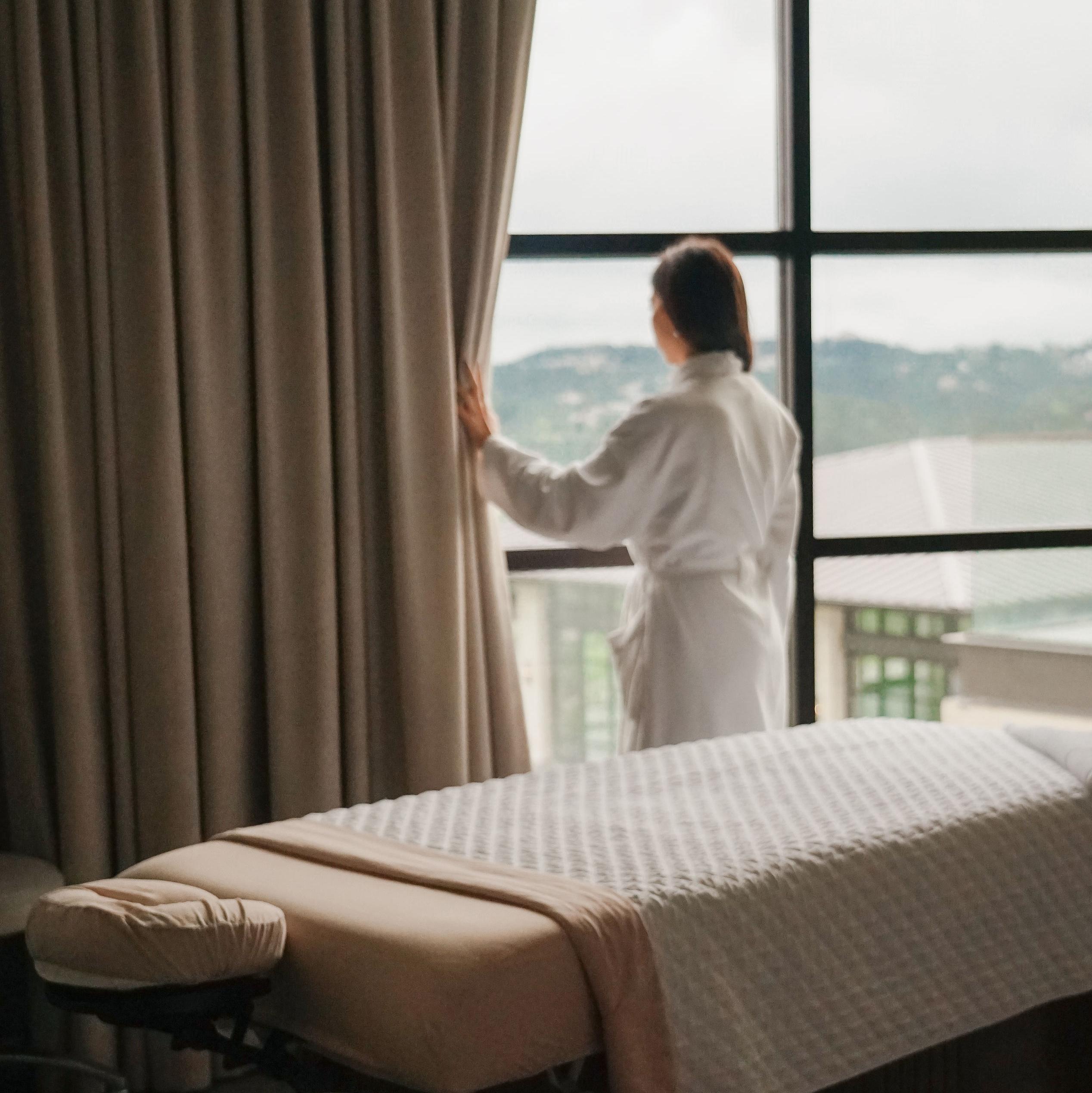 A robed woman looks out the window of the spa with a massage table behind her at Austin's Omni Barton Creek Resort & Spa 