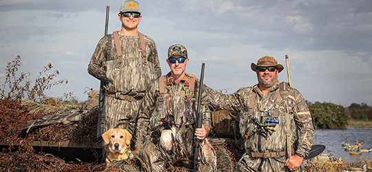 Three hunters in camouflage pose with a dog by the water.