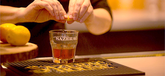 Hands squeeze a lemon peel over a Sazerac cocktail at Sazerac House in Louisiana.