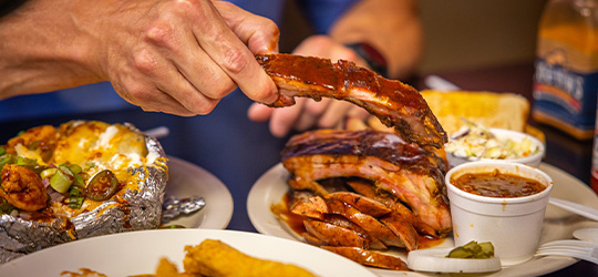 A hand reaches for perfectly sauced ribs, piled high on a plate with baked beans and other sides, surrounded by other classic barbecue dishes, like baked potatoes.