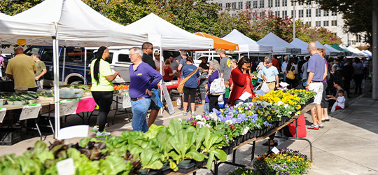 Shoppers stroll through a local farmers market in Louisiana, next to tents filled with local goods and tables filled with fresh flowers and greens.