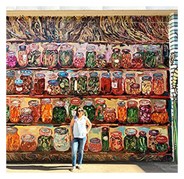 Woman standing outside in front of a large, colorful mural of various foods in jars