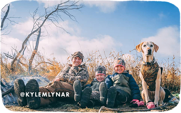 Man, two young girls and a dog sitting on the ground in a field wearing jackets and camouflage, with overlay text that reads: @kylemlynar