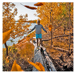 Man walking on a fallen tree log by a body of water, surrounded by trees with bright orange leaves