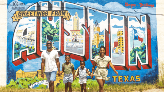 Two adults and two children walk while holding hands with the Greetings from Austin mural in the background