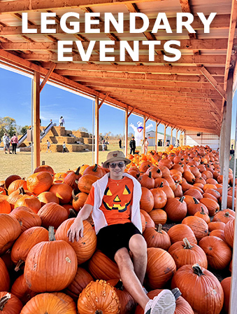 Man wearing a shirt with a jack-o-lantern on it, sitting on a large pile of orange pumpkins under an outdoor wooden structure, with overlay text that reads: Legendary Events