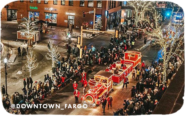 Aerial view of a holiday parade with holiday lights on a float, and overlay text that reads: @downtown_fargo