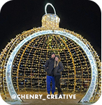 A man and a woman posing at night in a lit-up sculpture shaped like a round Christmas ornament, with overlay text that reads: @chenry_creative