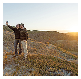 Two people standing on grassy hills taking a selfie with sunset in the background