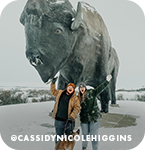 Two people and a dog posing in front of a large buffalo sculpture on a snowy day, with overlay text that reads: @cassidynicolehiggins