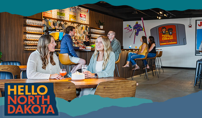 Two women sitting at a wood table in a bar having a drink, with a people sitting at a bar top behind them, with overlay text that reads: Hello North Dakota