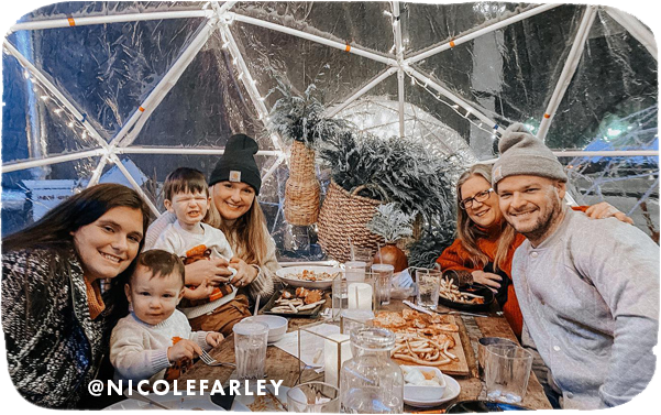 A group of three women, a man and three young boys eating at a table inside a plastic dome with holiday lights on it, with overlay text that reads: @nicolefarley