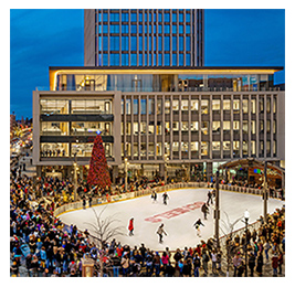 Aerial view of an outdoor ice skating rink at night, surrounded by lit-up buildings and a crowd of people