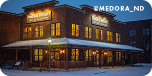 Brick building at night with illuminated windows on a snowy street, with overlay text that reads: @medora_nd