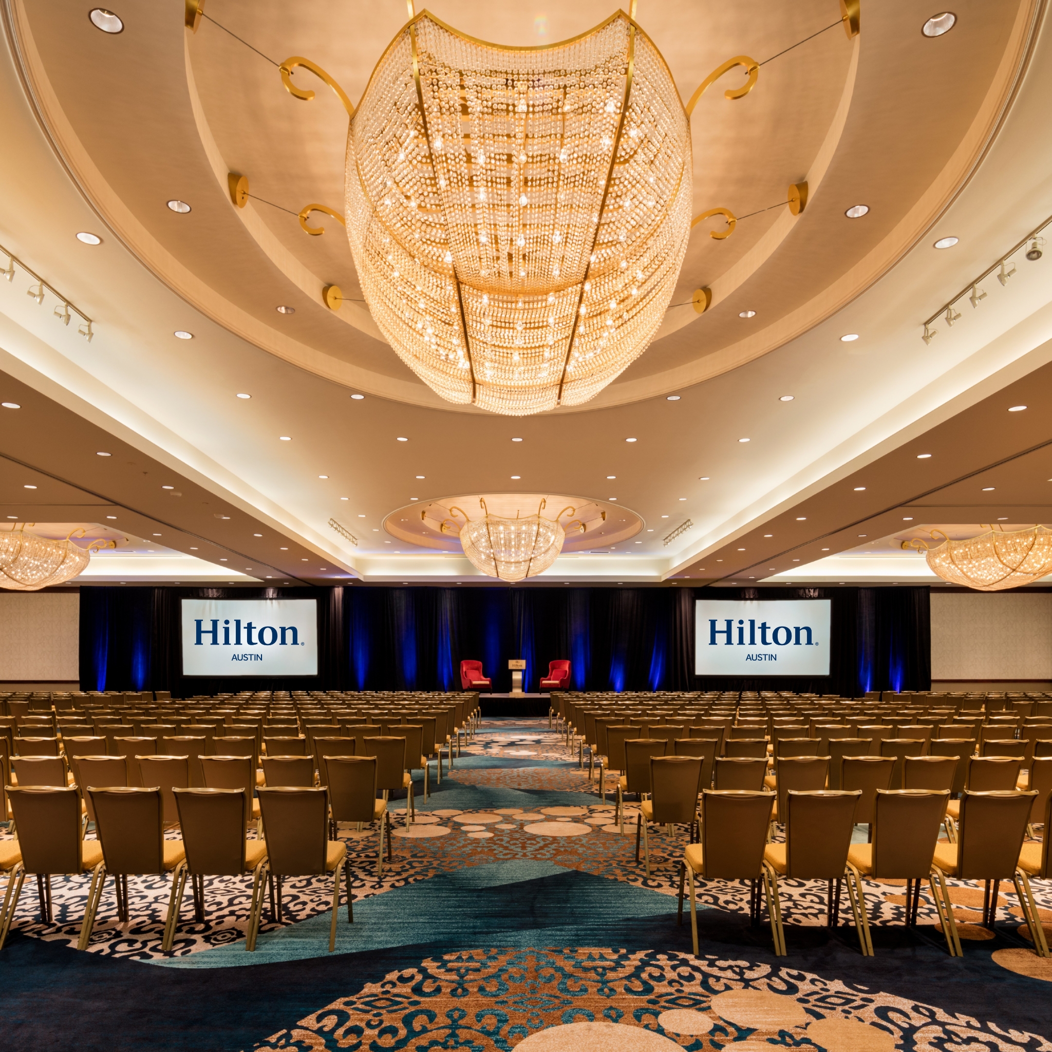 The fourth-floor ballroom at the Hilton Austin with rows of chairs facing a stage holding a podium and two red wingback chairs, all under a gold-hued ceiling and elaborate light fixtures.