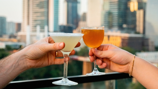 Two hands, each holding a cocktail, toast on the rooftop of The Loren Hotel Austin, with the city skyline in the background