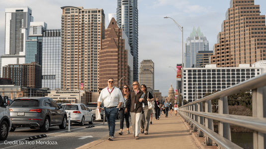 People walk on a pedestrian bridge crossing Lady Bird Lake with the Austin city skyline behind them